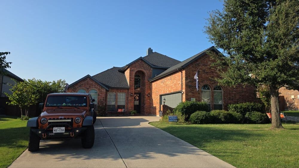 Brick house with car in driveway, green lawn, and trees under clear blue sky.